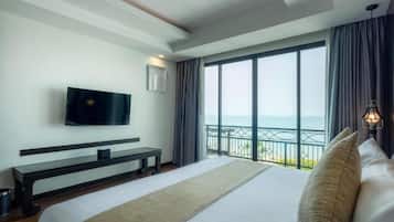 A hotel room with a large bed, a flat-screen TV, a bench, a balcony with a view of the sea, and a nightstand with a lamp.