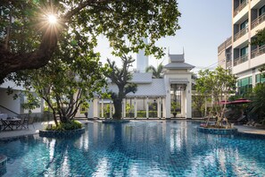 A swimming pool with a tiled edge, surrounded by trees and a building with a white facade and a tiled roof.