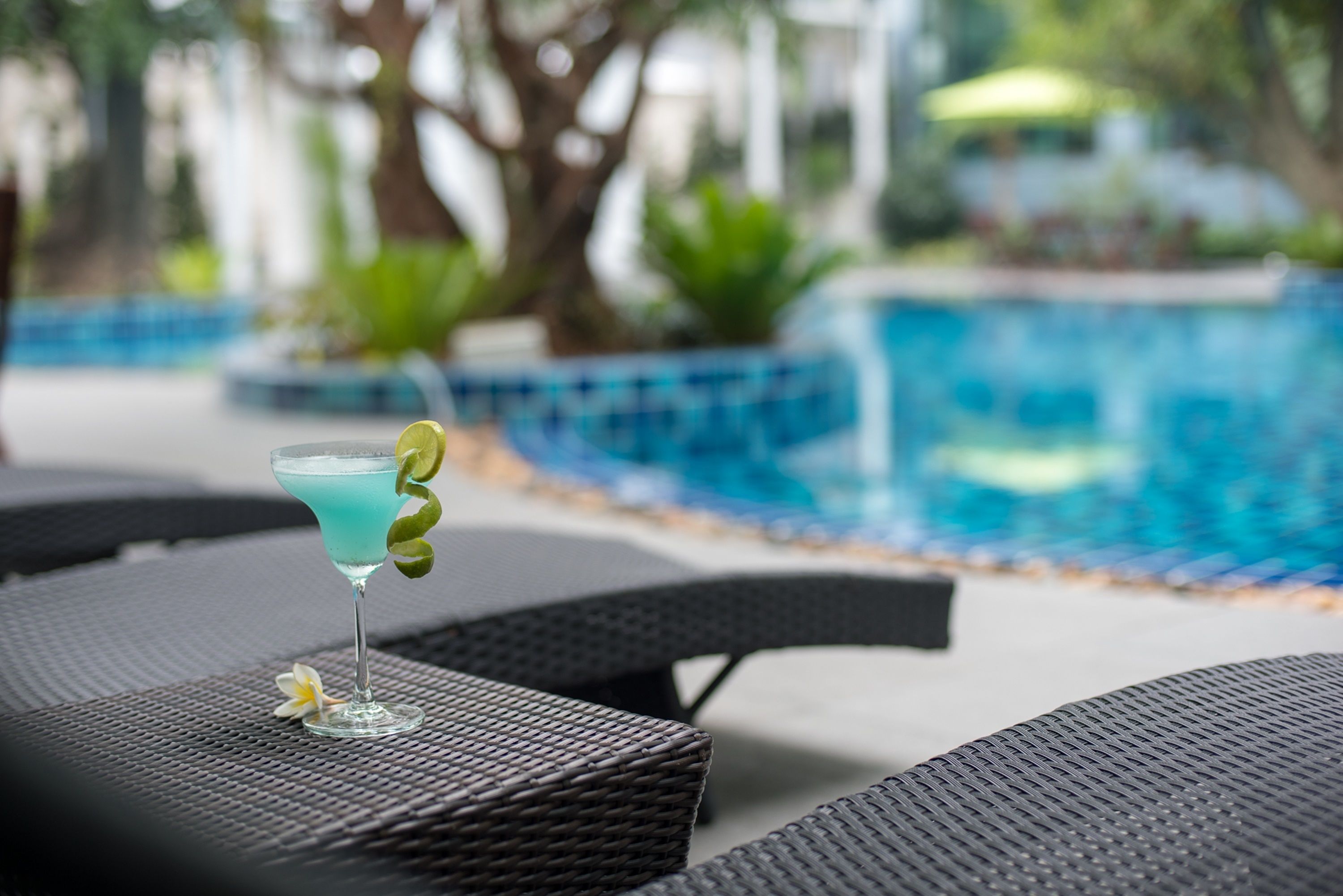 A cocktail on a wicker table near a pool with a blurred background of greenery and a poolside area.