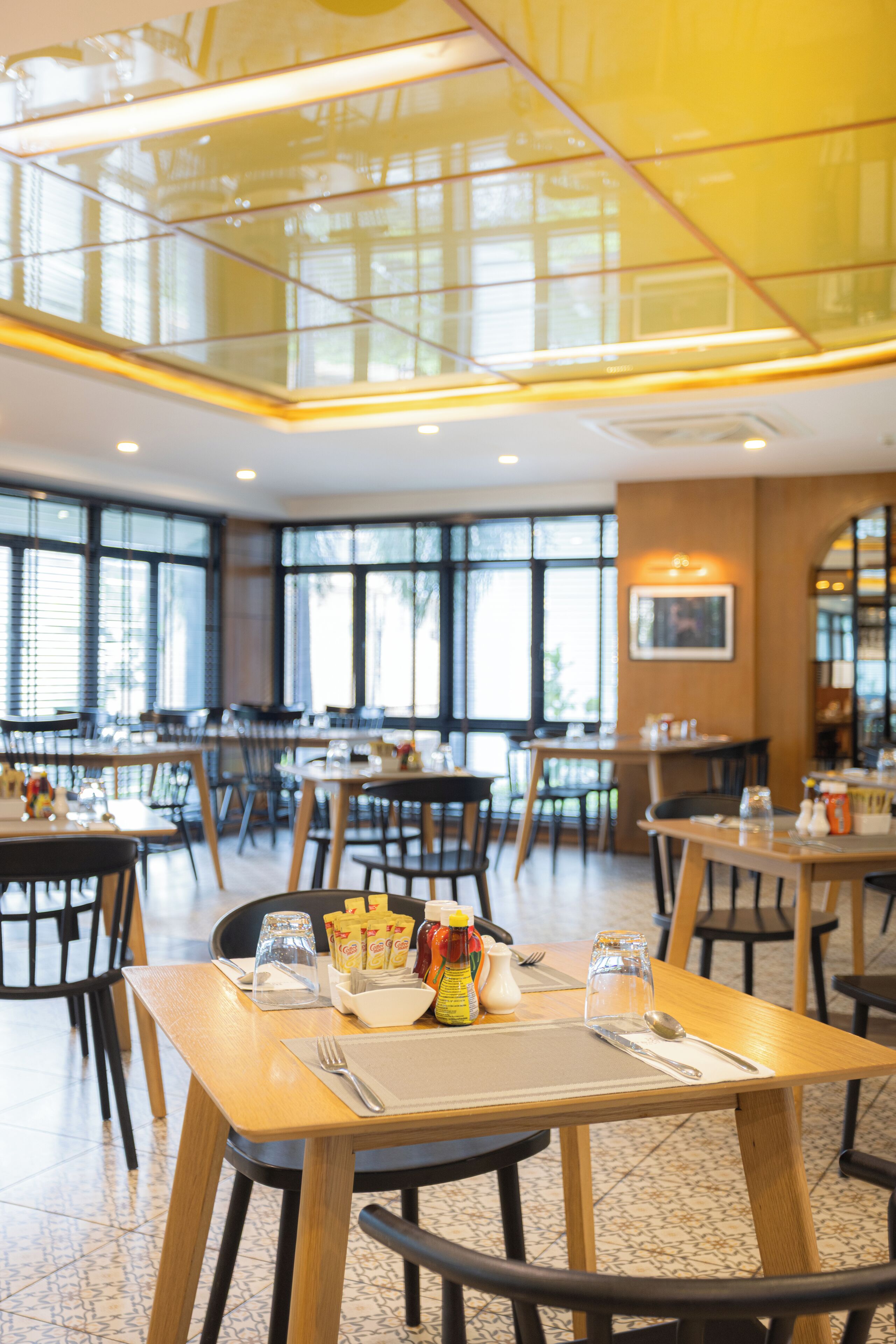 A spacious restaurant with high ceilings and large windows, featuring wooden tables and chairs, and a variety of condiments and utensils on the tables.