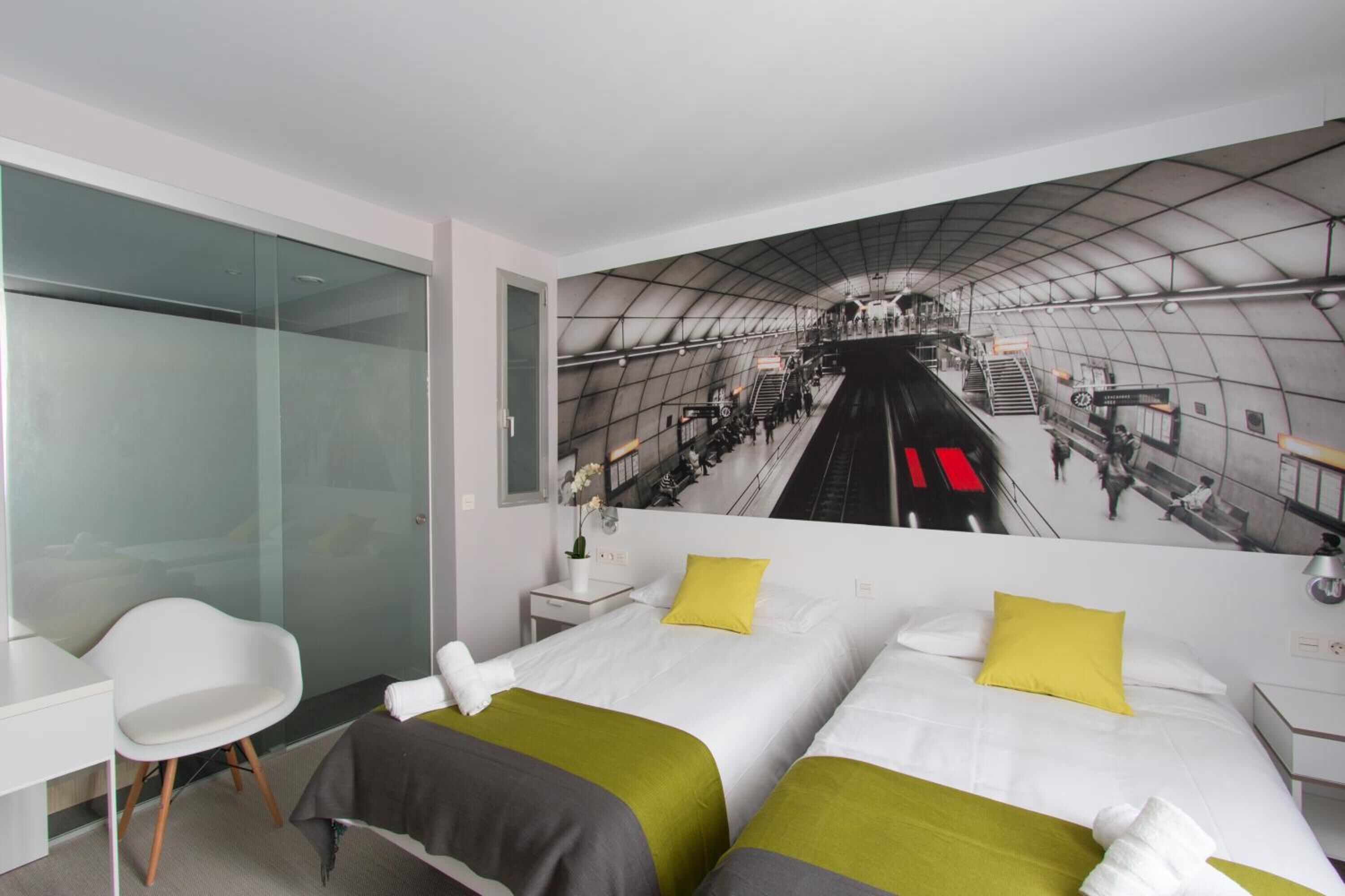 Habitación de hotel con dos camas, un separador de vidrio, un escritorio blanco y un mural de una estación de tren.