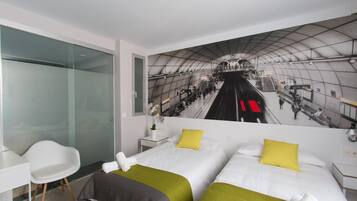 Habitación de hotel con dos camas, un separador de vidrio, un escritorio blanco y un mural de una estación de tren.