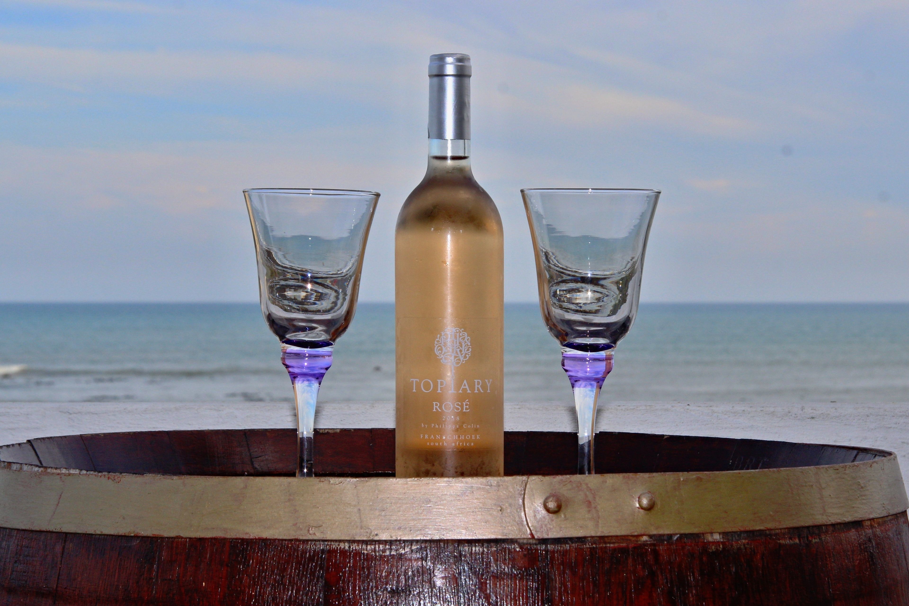 A bottle of Topiary Rosé and two glasses on a wooden barrel by the beach.