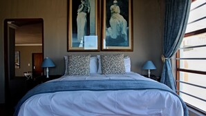 Una habitación de hotel con una cama grande, dos cuadros en la pared, una ventana con cortinas y una mesita de noche con una lámpara.