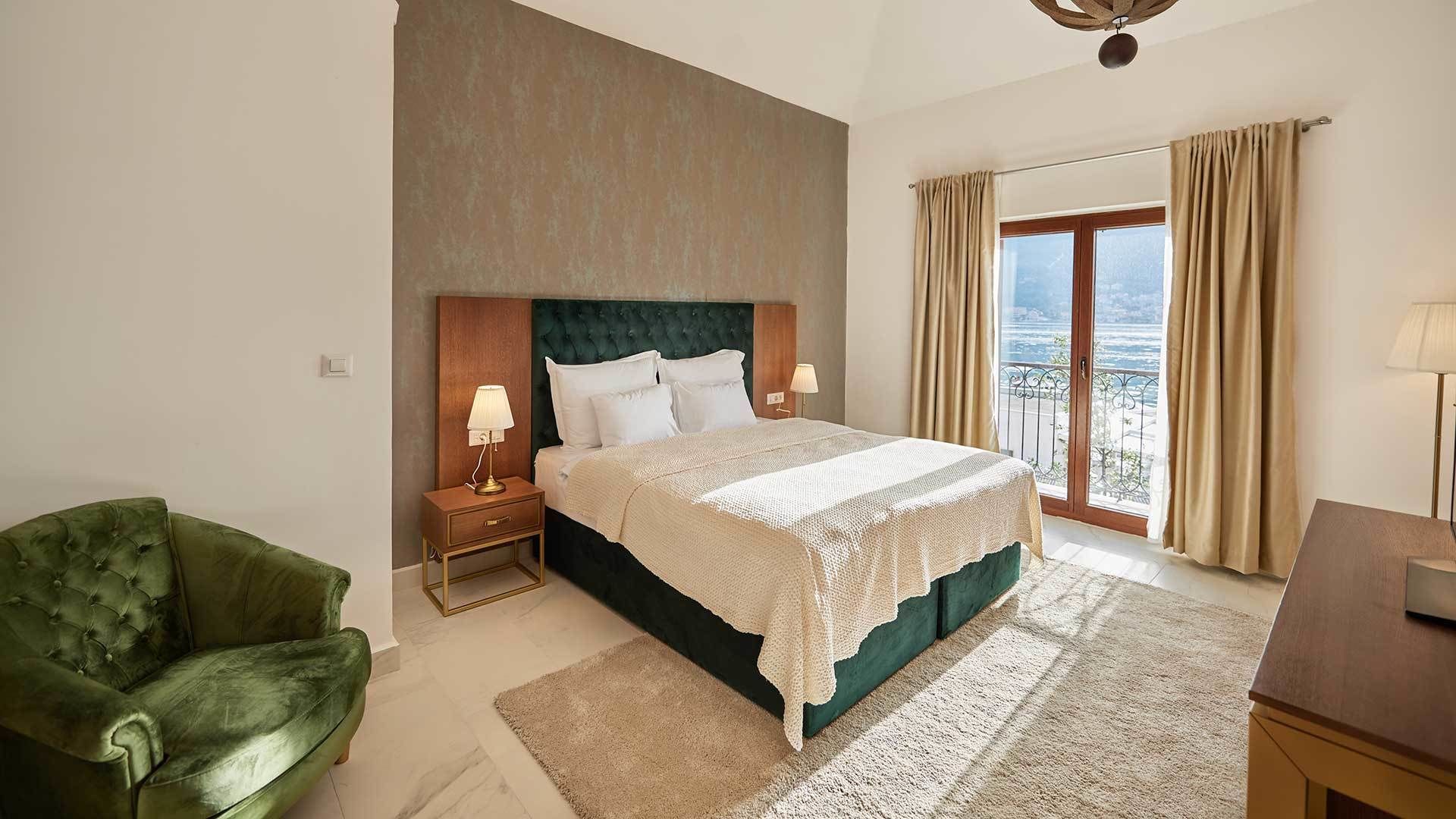 A bedroom with a large bed, a green armchair, a nightstand with lamps, and a view of the sea through a balcony.