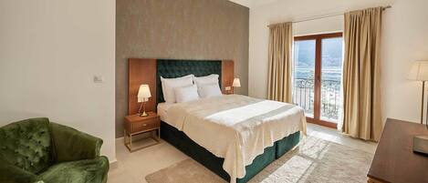 A bedroom with a large bed, a green armchair, a nightstand with lamps, and a view of the sea through a balcony.