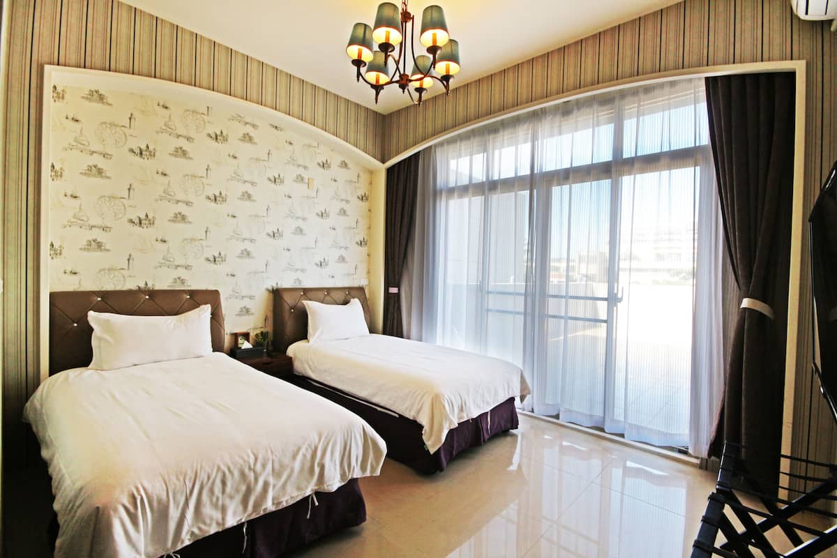 A hotel room with two beds, a large window with curtains, a chandelier, and a wall with a pattern.