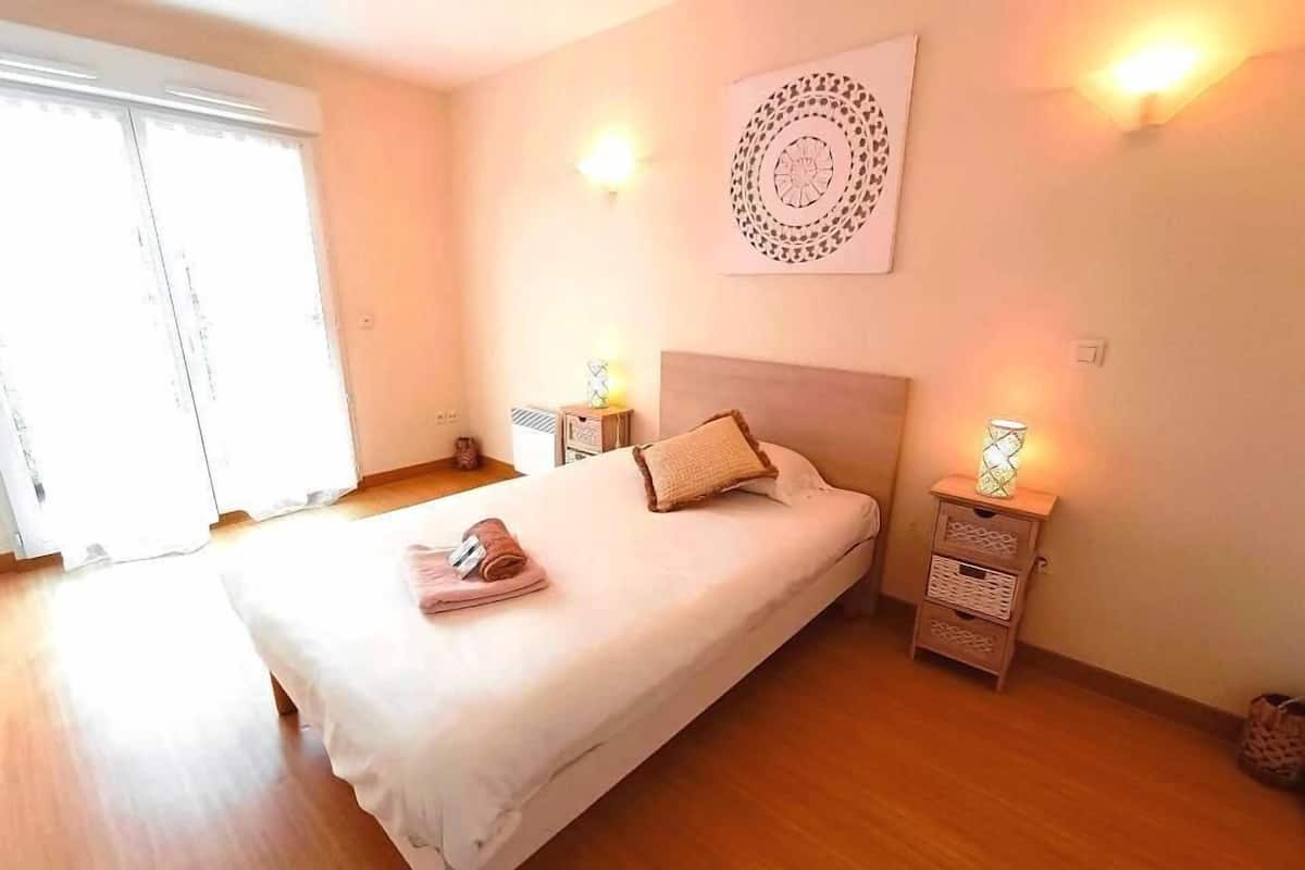 Una cama bien hecha con edredón blanco, una toalla marrón y una sola almohada. Hay dos mesitas de noche con lámparas, un adorno en la pared y un ventanal con persianas.