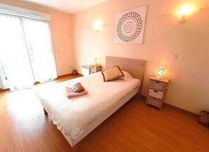 A neatly made bed with a white duvet, a brown towel, and a single pillow. There are two bedside tables with lamps, a wall-mounted decorative item, and a large window with blinds. - Résidence Cintray Parc (Breteuil-sur-Iton)
