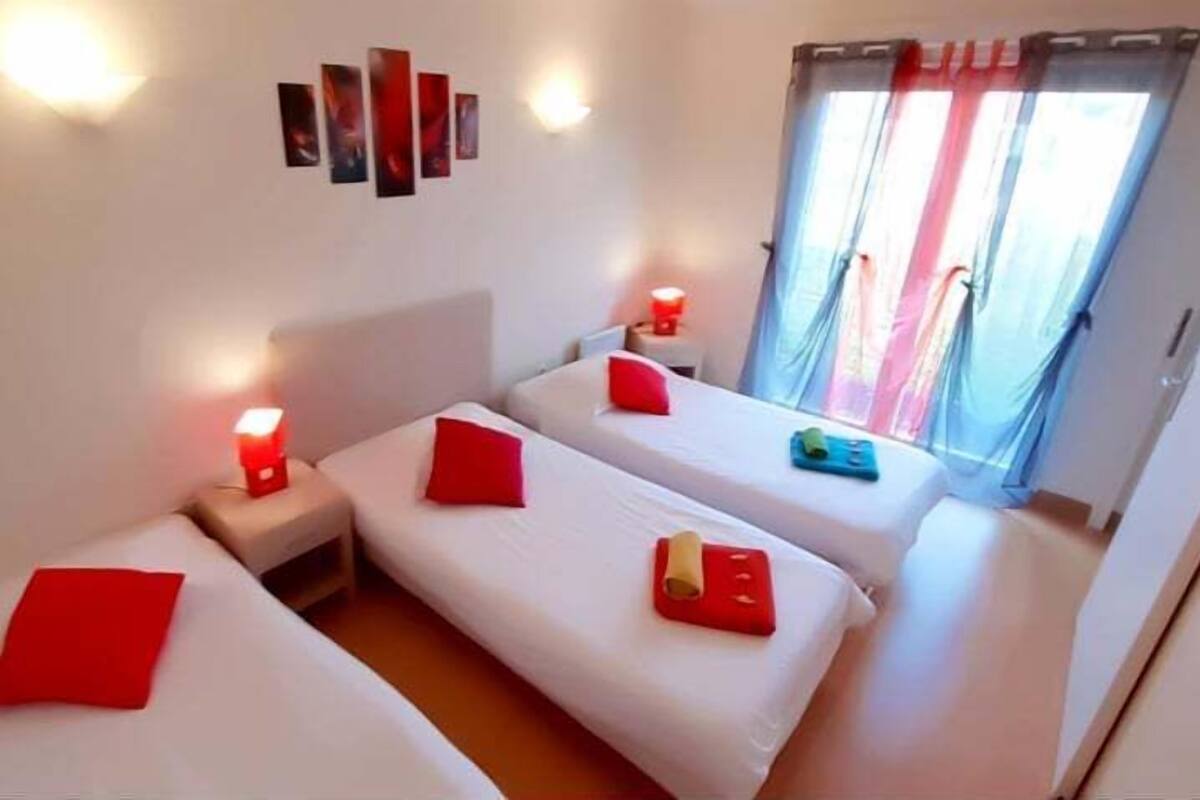 Una habitación con tres camas individuales, cada una con un almohadón rojo y una mesita de noche con una lámpara roja. Hay luces fijadas en la pared y un ventanal con cortinas.
