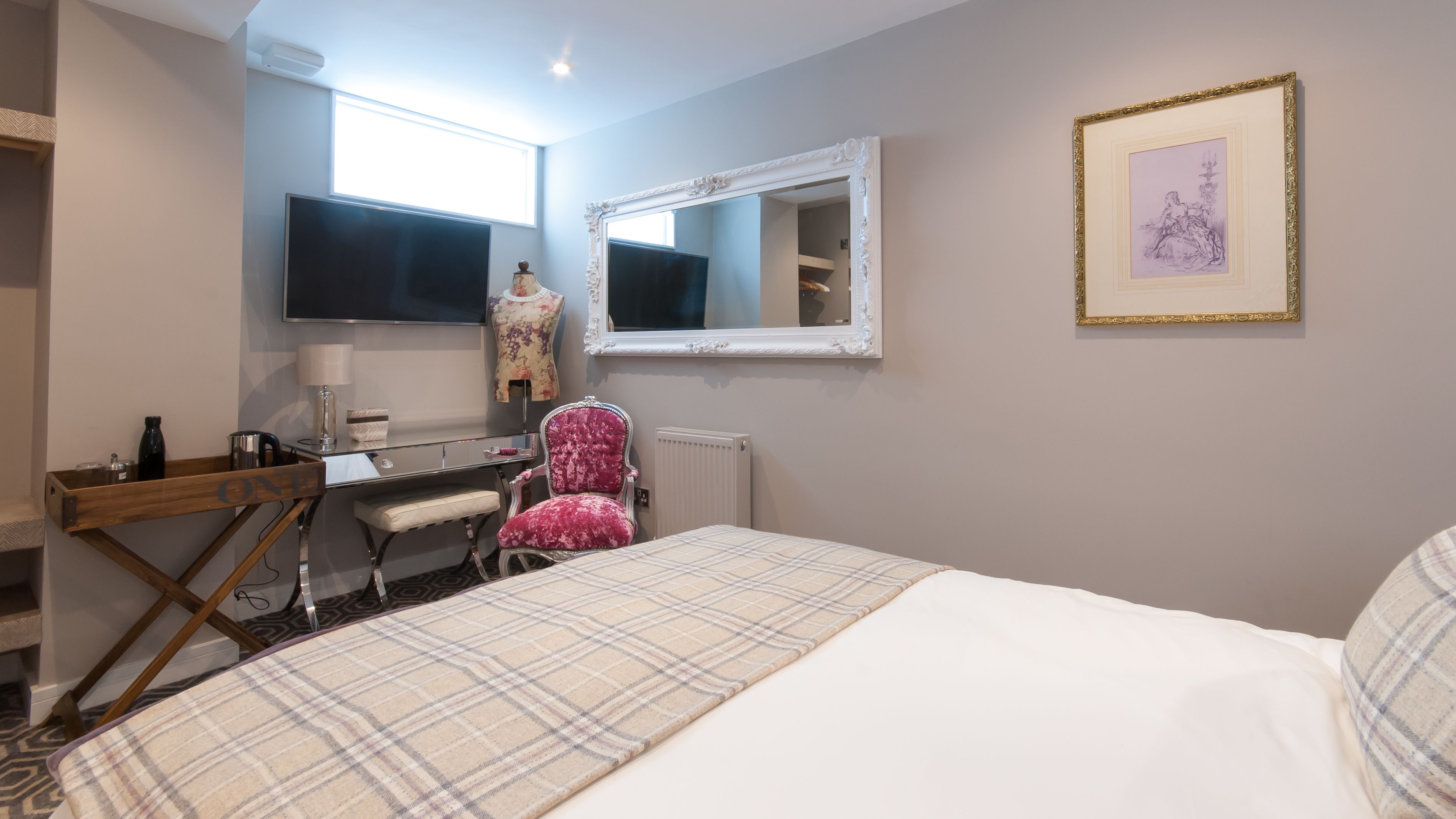 Hotellihuone, jossa on sänky, työpöytä tietokoneella, tuoli, suuri peili ja seinällä kehystetty taulu.