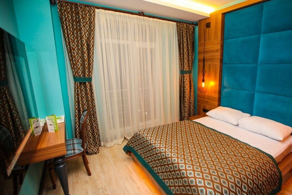 A bedroom with a bed, a desk, and a chair. There is a window with curtains and a wall-mounted light fixture.
