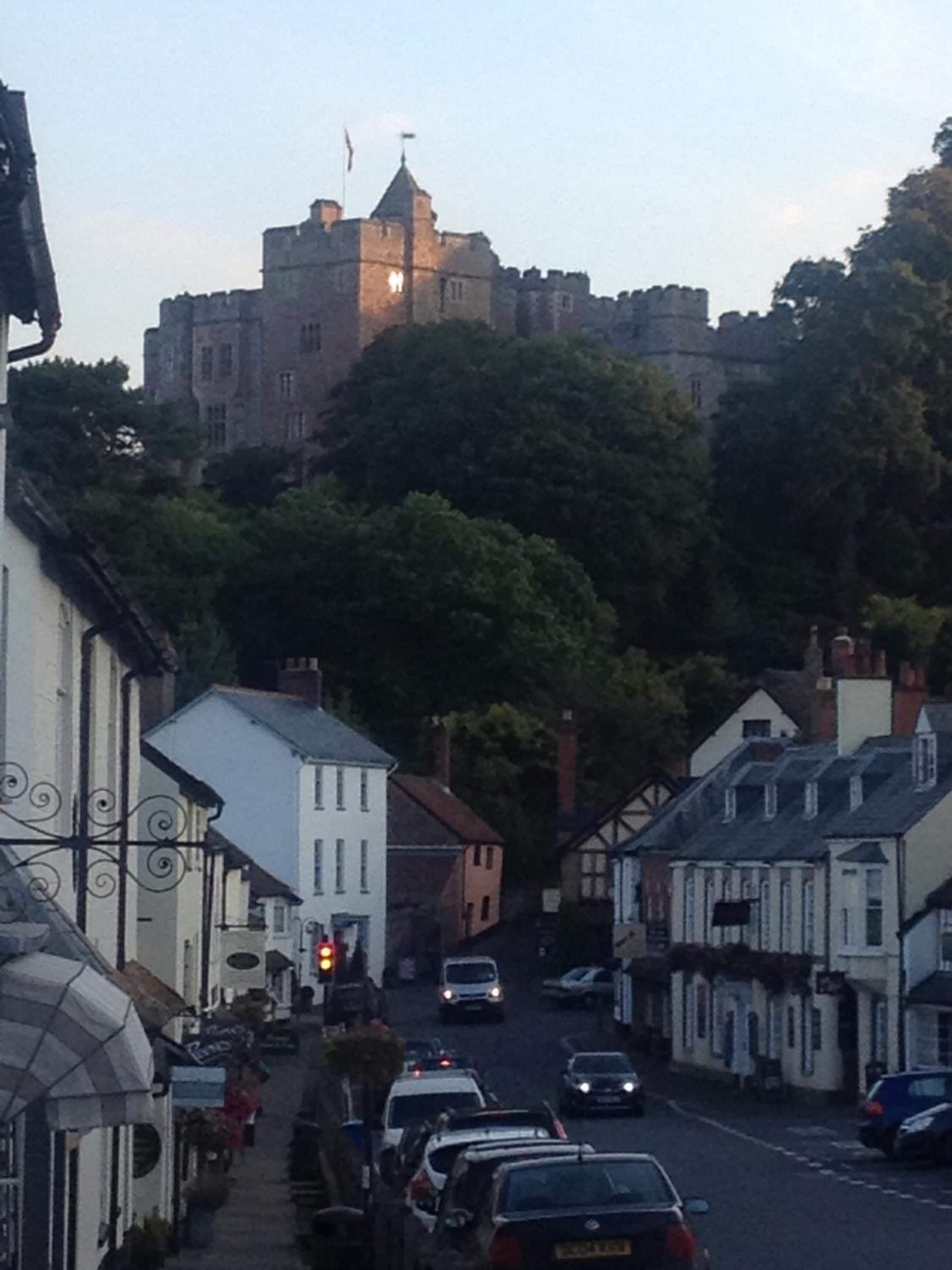 A street leading to a castle-like building with a prominent tower, surrounded by traditional buildings and parked cars.