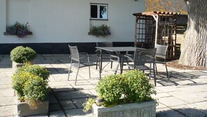 An outdoor patio with a table and chairs, potted plants, and a building with a window and flower boxes.