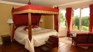A four-poster canopy bed with a wooden frame and red and white bedding, a small table with a chair, and a view of greenery through the window.
