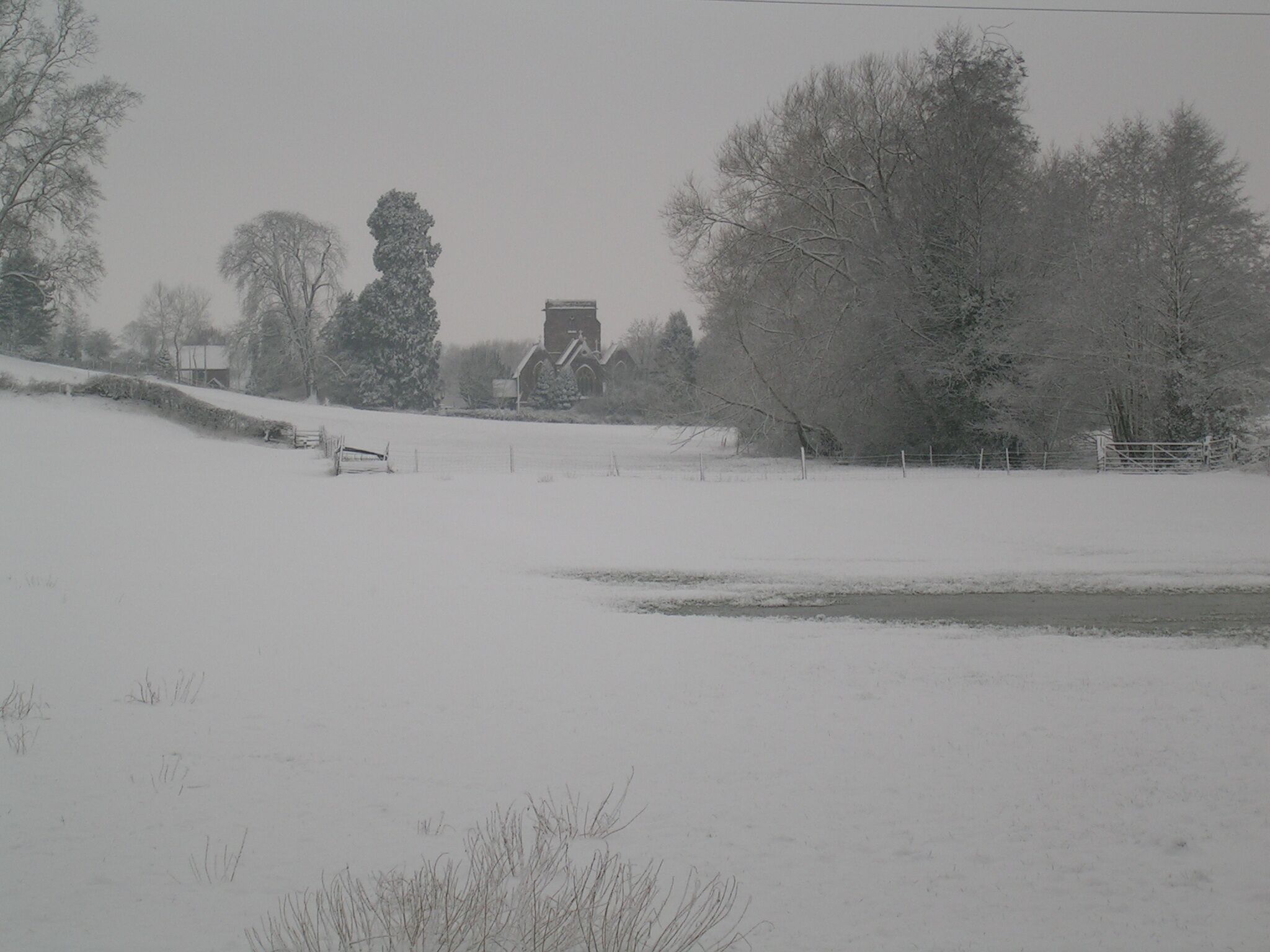 A snow-covered landscape with a building in the distance, surrounded by trees and a fence.