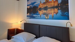 A hotel room with a bed, a nightstand, a lamp, and a large framed picture of a historic building reflected in a body of water. - Leine Hotel (Pattensen)