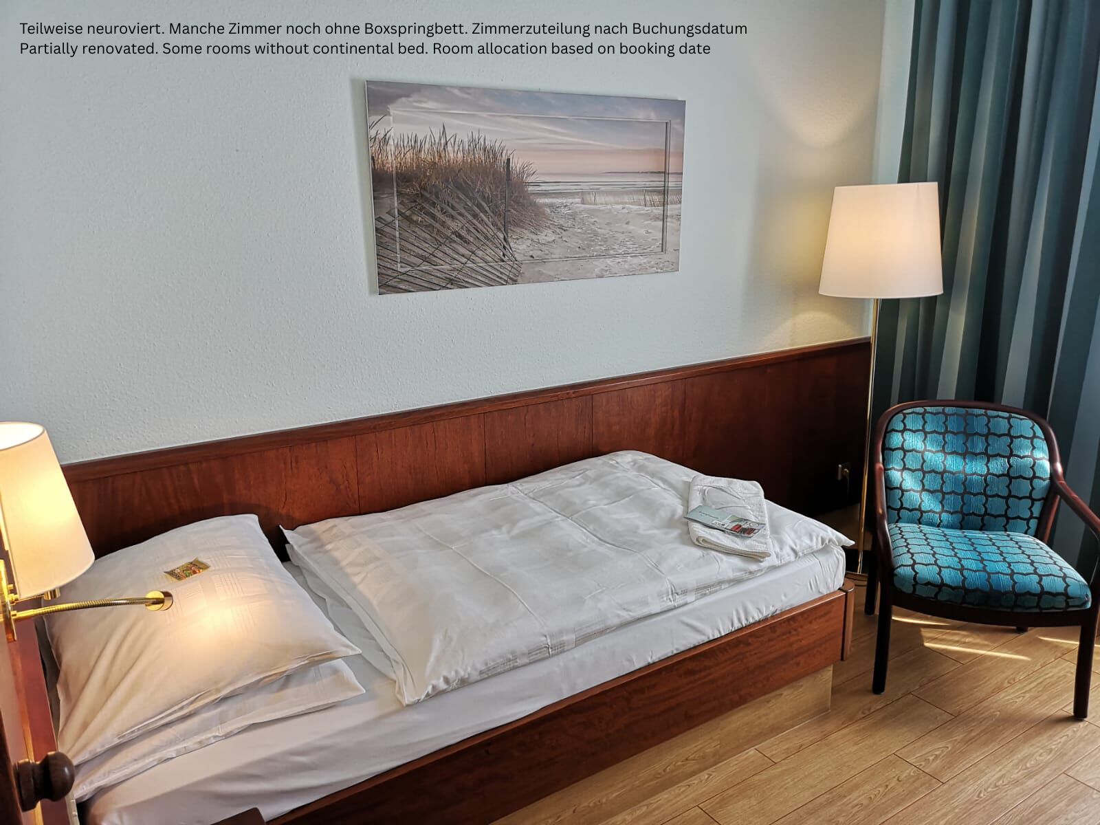 Een gedeeltelijk gerenoveerde kamer met een houten hoofdeinde, een enkel bed met witte lakens, een blauw gestreept fauteuil en een wandlamp.