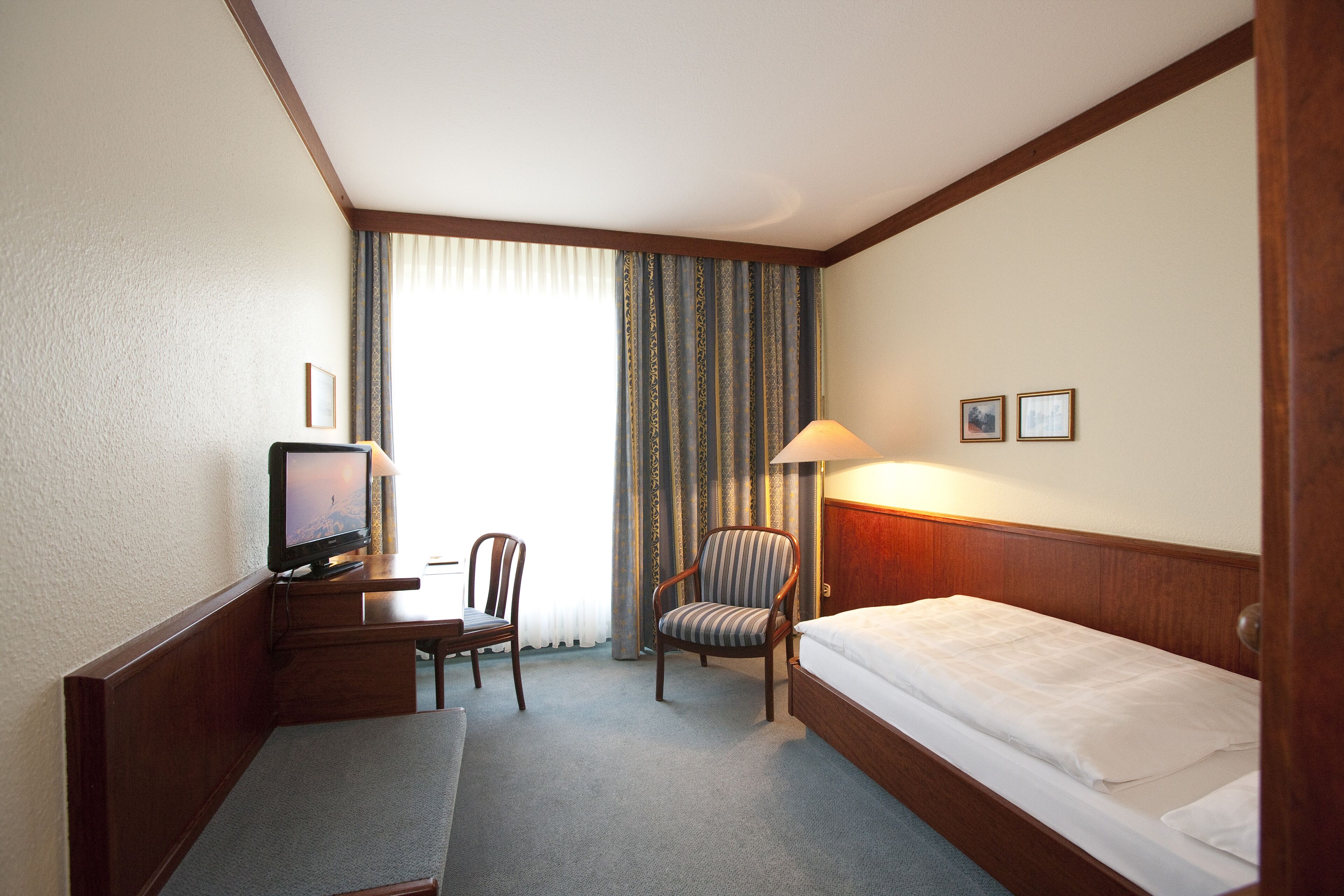 A hotel room with a bed, a desk with a television, a chair, and a lamp.