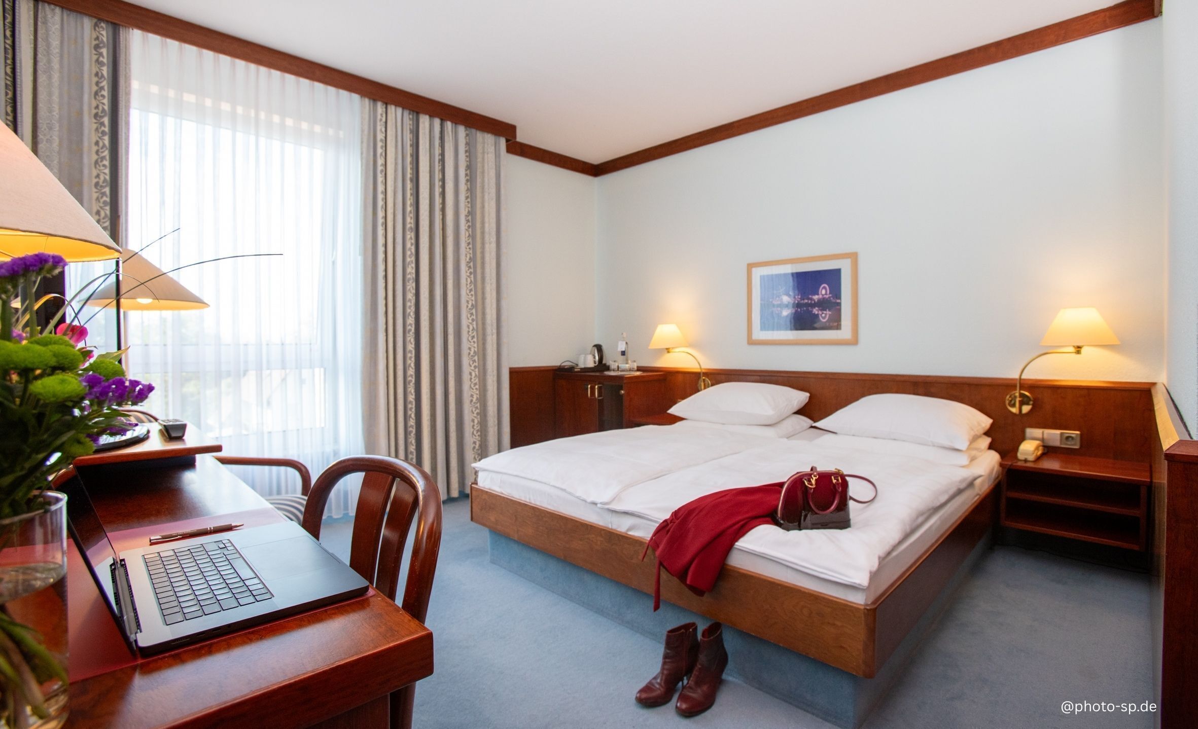 Een hotelkamer met een bed, een bureau met een laptop, een stoel, een lamp en een foto aan de muur.