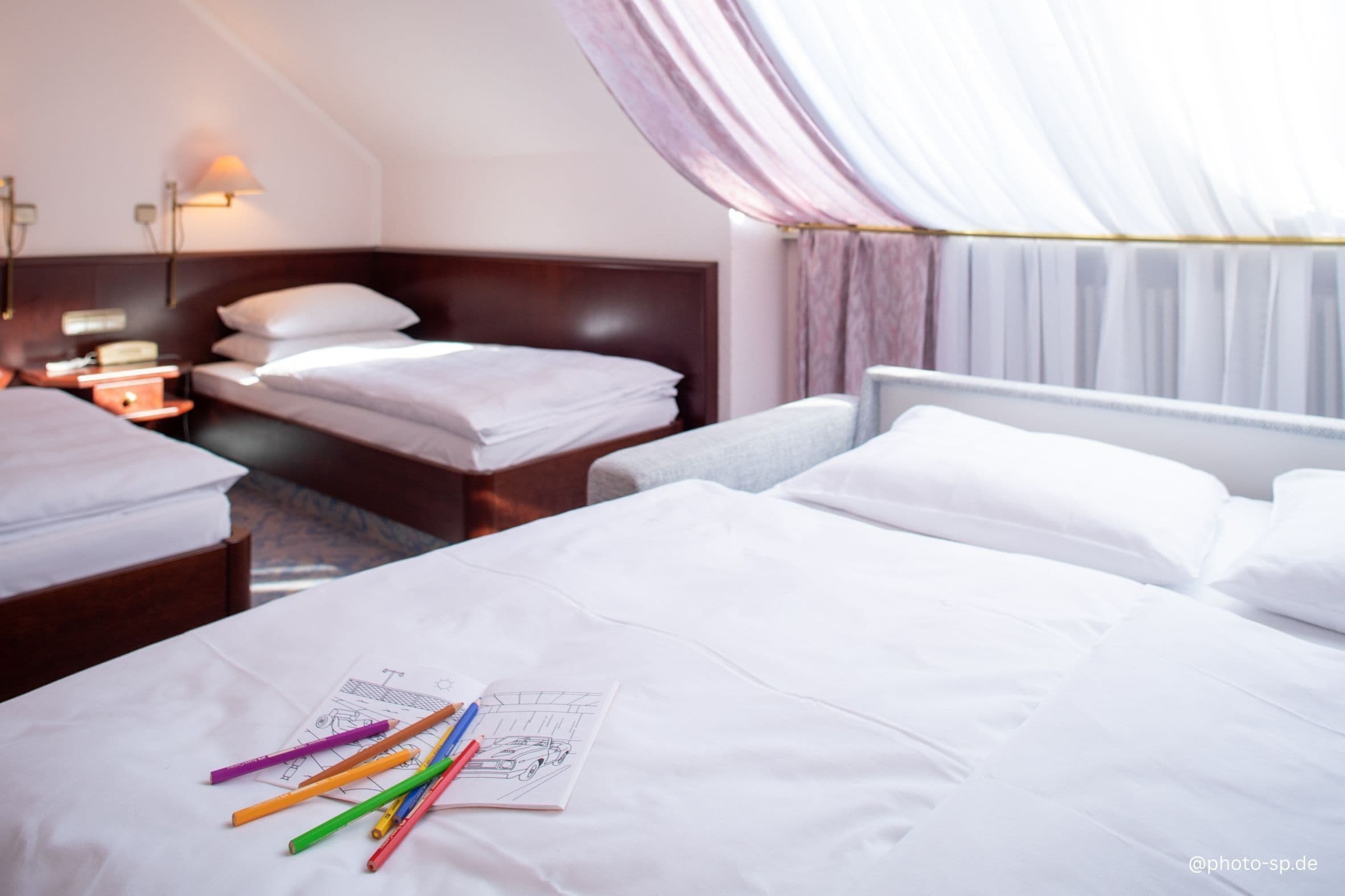 Camera d'albergo con due letti, un comodino e un tavolo con matite colorate e un disegno.