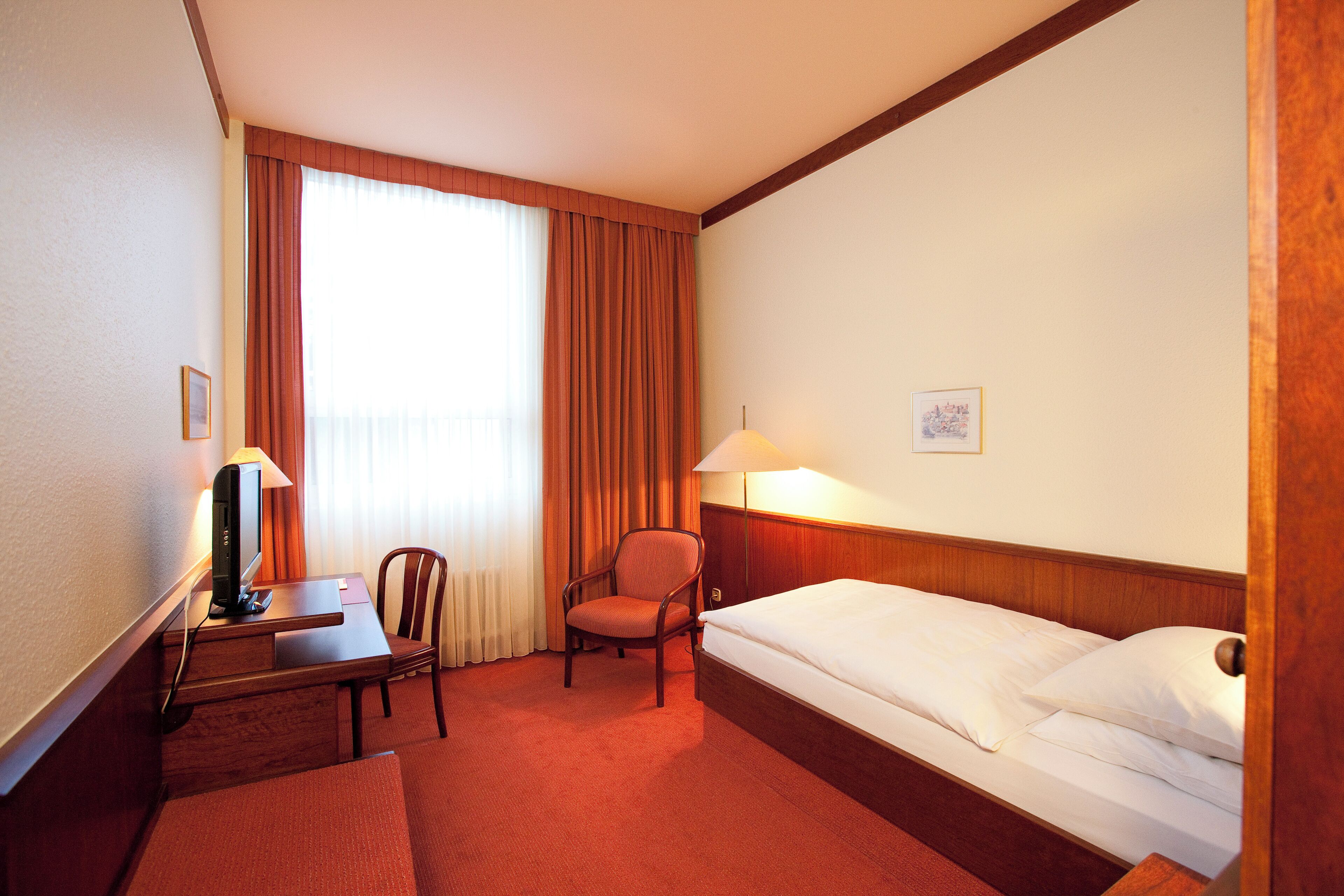 A hotel room with a bed, desk, chair, and a lamp.
