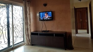 Una habitación con televisión montada en la pared, un mueble de madera oscura, un ventanal grande con rejas decorativas y una puerta que da a otra habitación.