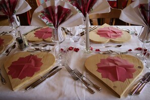 Una mesa dispuesta con platos de madera con forma de corazón, cubiertos y copas, decorada con flores de papel rosa y pétalos de rosa.