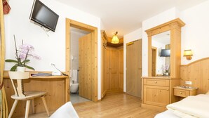 A room with a wooden desk, chair, and a bed. There is a television mounted on the wall and a mirror with a lamp. - Hotel Alpenrose (La Valle)