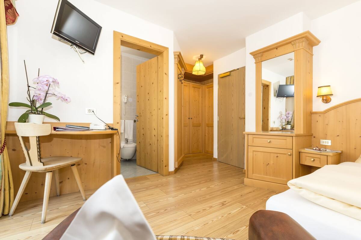 Een kamer met een houten bureau, een stoel en een bed. Er is een televisie aan de muur bevestigd en een spiegel met een lamp.