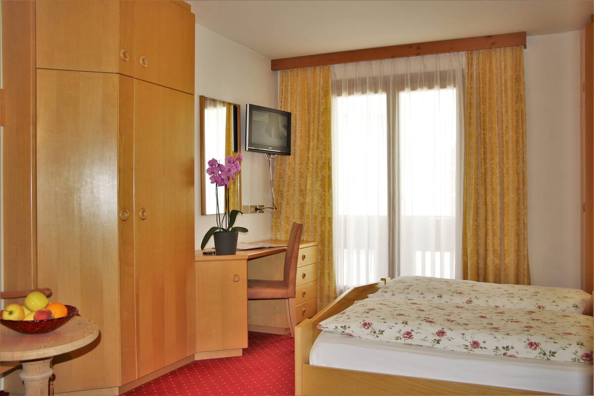 Hotelkamer met een houten bureau, een stoel, een bed met bloemenprint linnen en een televisie, plus een raam met gordijnen.