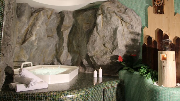 A spa area with a large rock wall, a hot tub, towels, candles, and a wooden clock. - Hotel Alpenrose (La Valle)