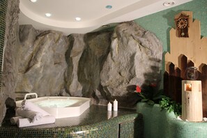 Un área de spa con una gran pared de roca, una bañera de hidromasaje, toallas, velas y un reloj de madera.