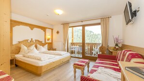 A bedroom with a wooden bed, a sofa, a table, and a TV. - Hotel Alpenrose (La Valle)