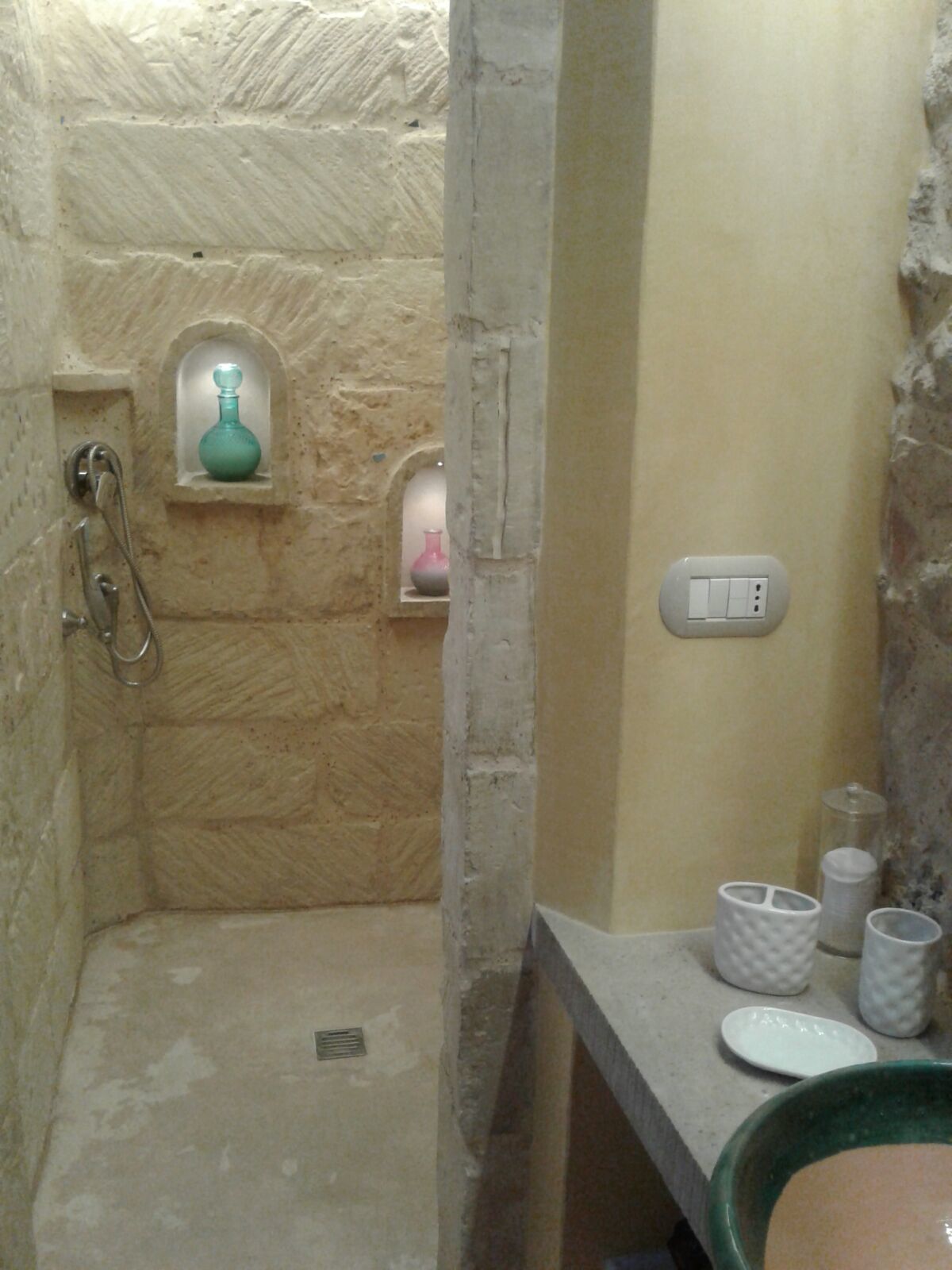 Une salle de bain avec un mur en pierre, une douche, un lavabo avec des articles de toilette et un interrupteur.