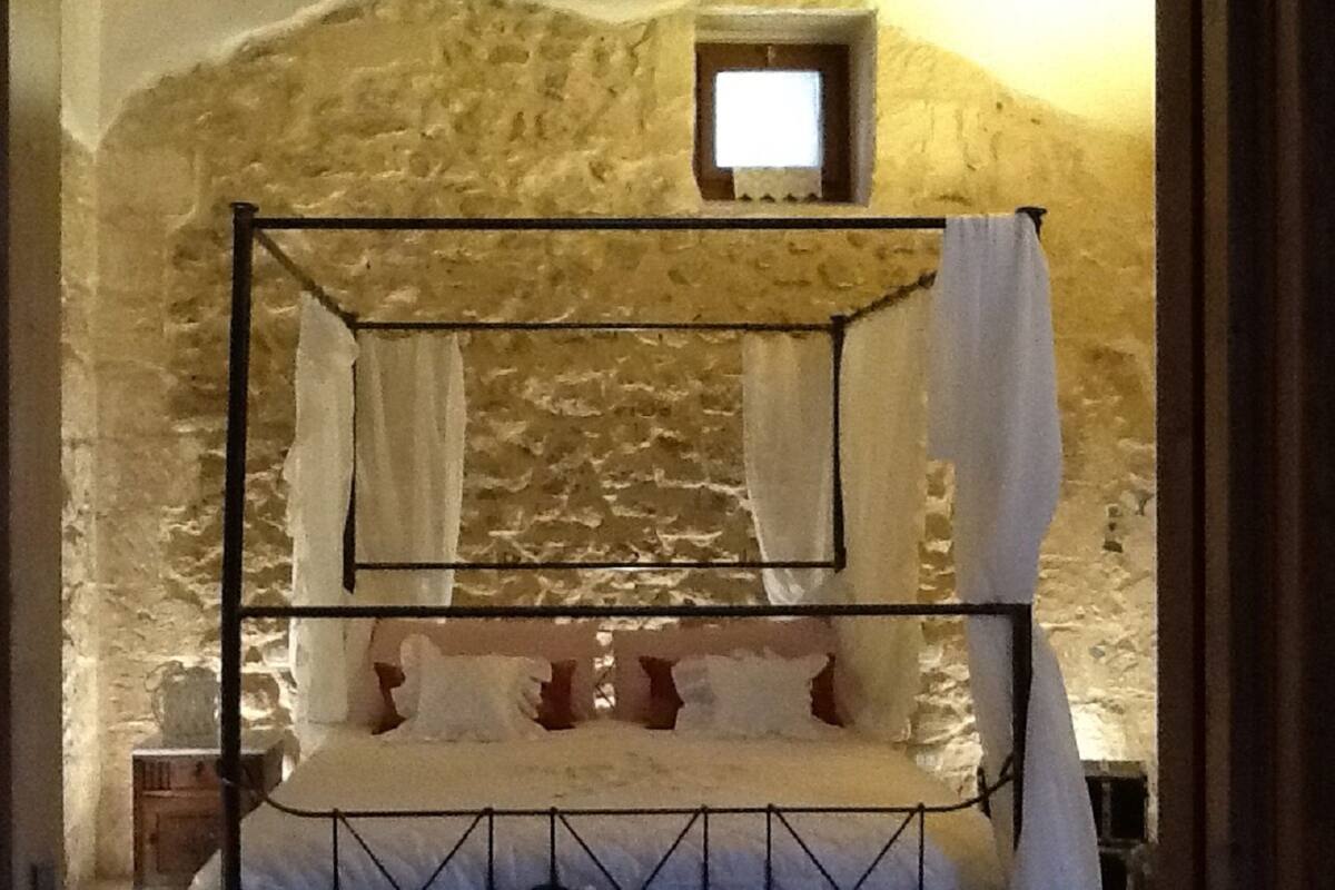 Un lit à baldaquin avec du linge de lit blanc et des rideaux, placé contre un mur de pierre avec une petite fenêtre au-dessus.