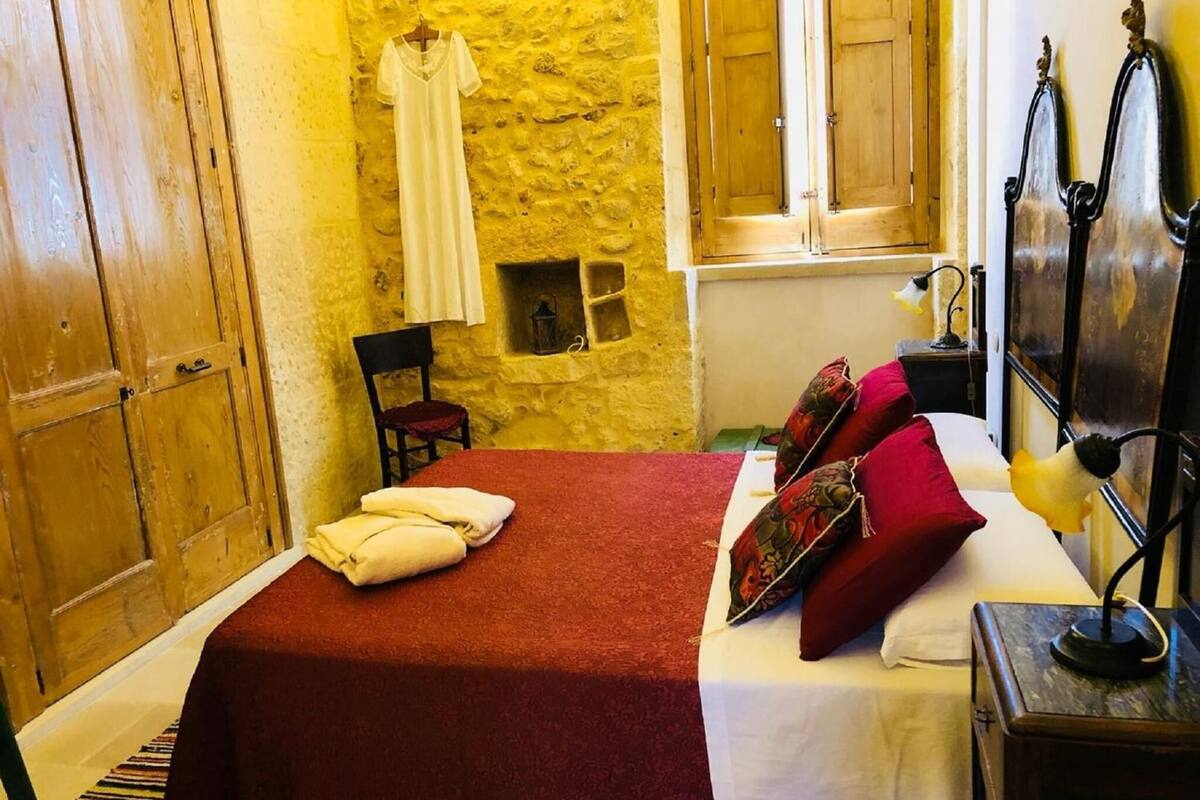 Une chambre avec un mur en pierre, une armoire en bois, un lit avec une literie rouge, des oreillers et une table de chevet avec une lampe.