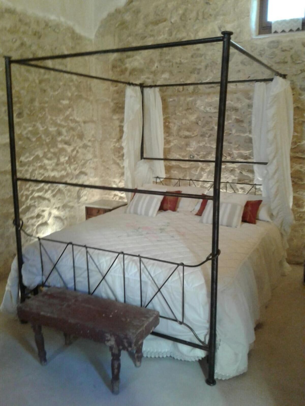 A four-poster bed with a canopy, a wooden bench, and a stone wall background.