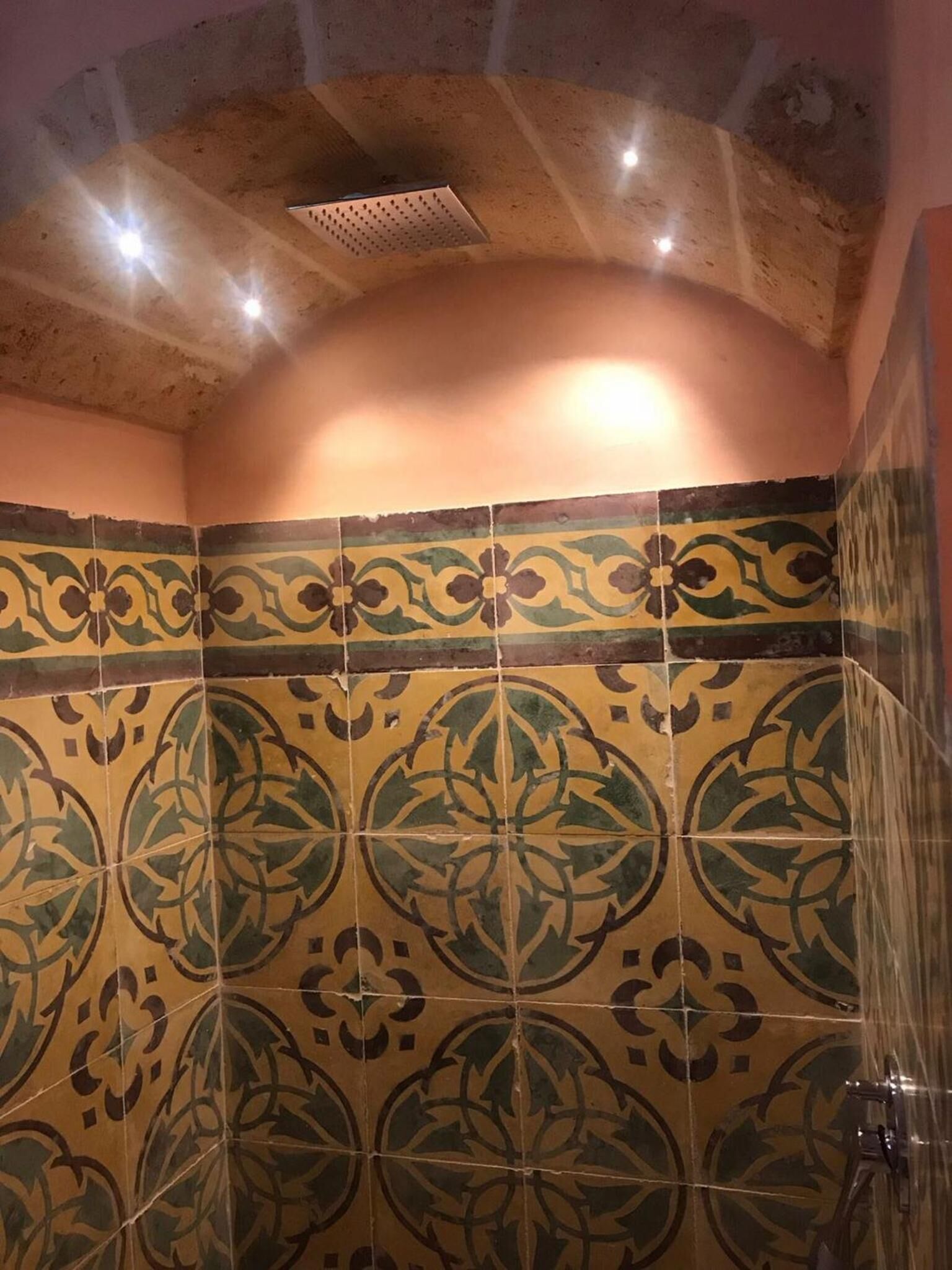 Une salle de bain aux carreaux verts et jaunes décorés, avec une douche et un plafond laissant apparaître la maçonnerie.
