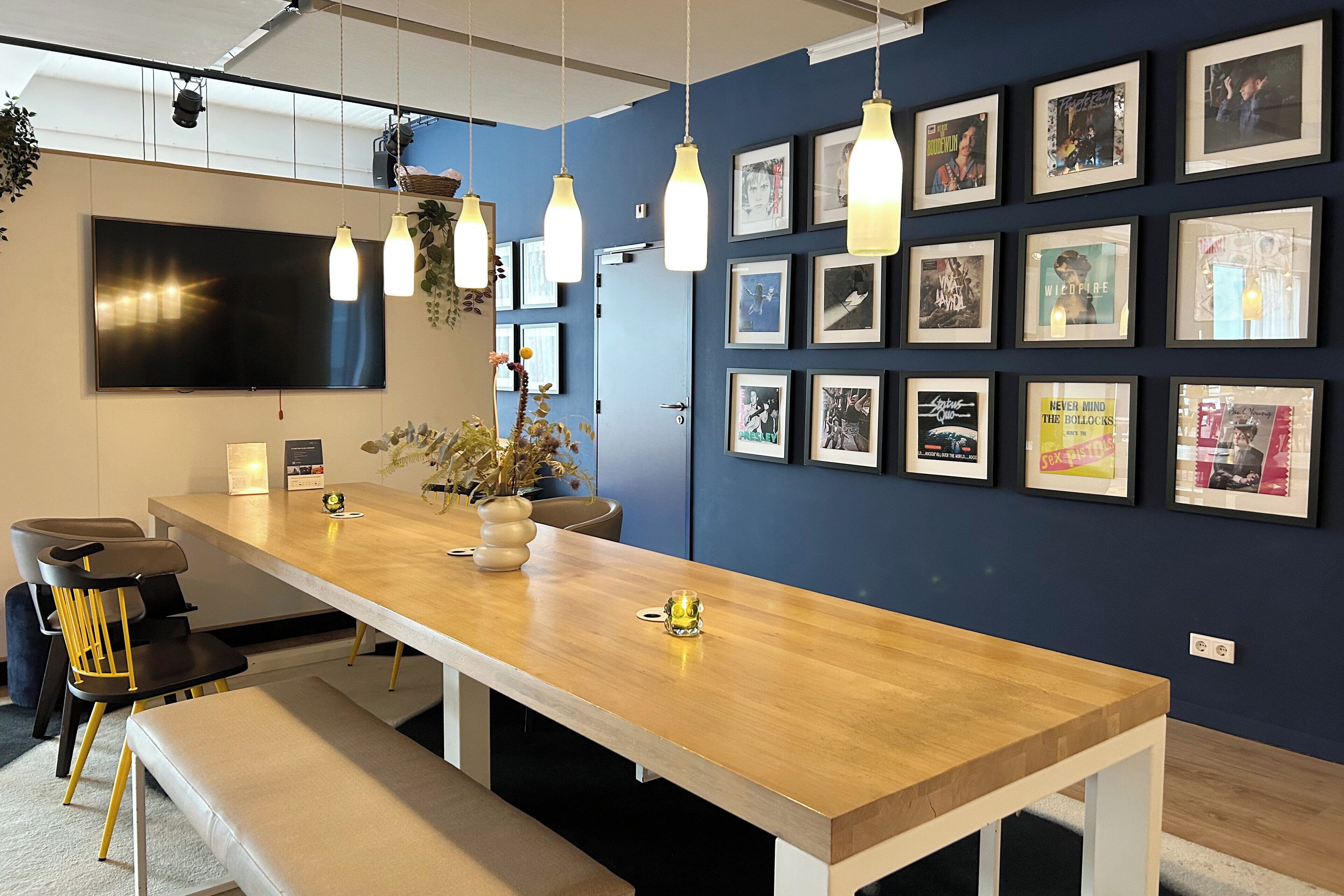 A modern office space with a long wooden table, multiple chairs, and a large flat-screen TV mounted on the wall.