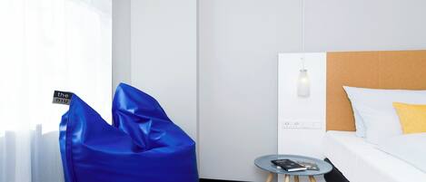 A hotel room with a bed, a bedside table with a book, a small round table, and a blue bean bag chair.
