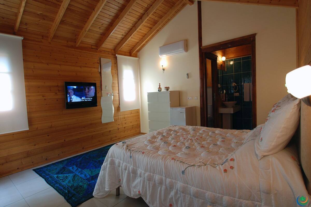 A bedroom with a wooden ceiling, a bed with a floral duvet, a television on the wall, and a small white dresser.