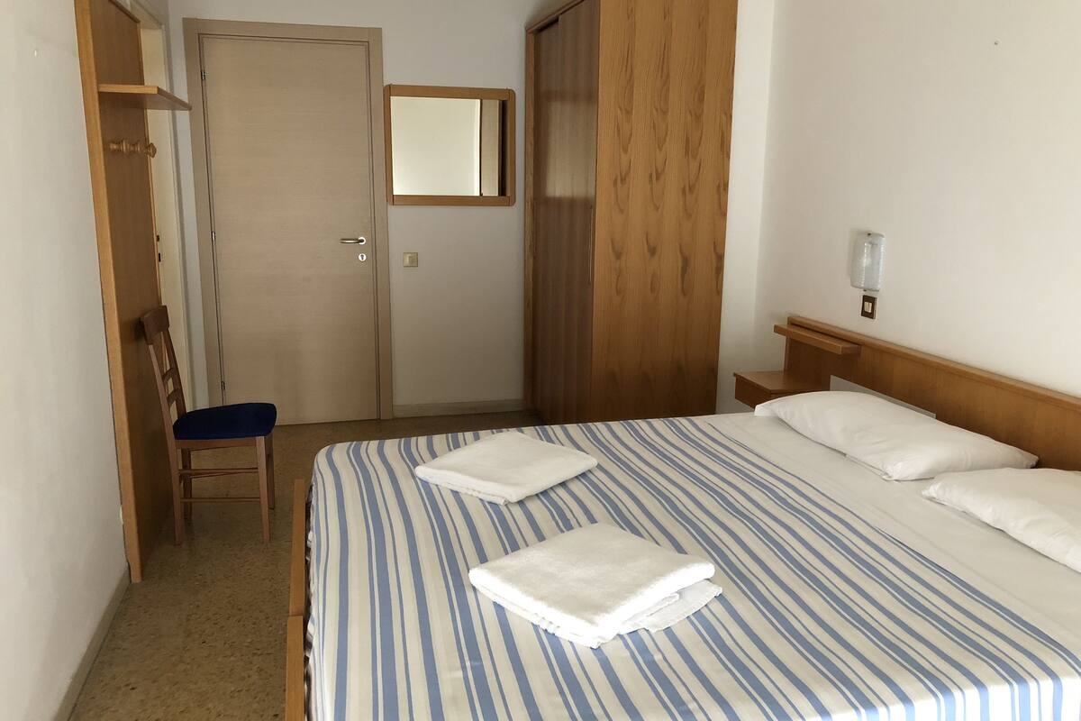 Une chambre d’hôtel avec un lit, une chaise, une armoire, un miroir et une table de chevet.