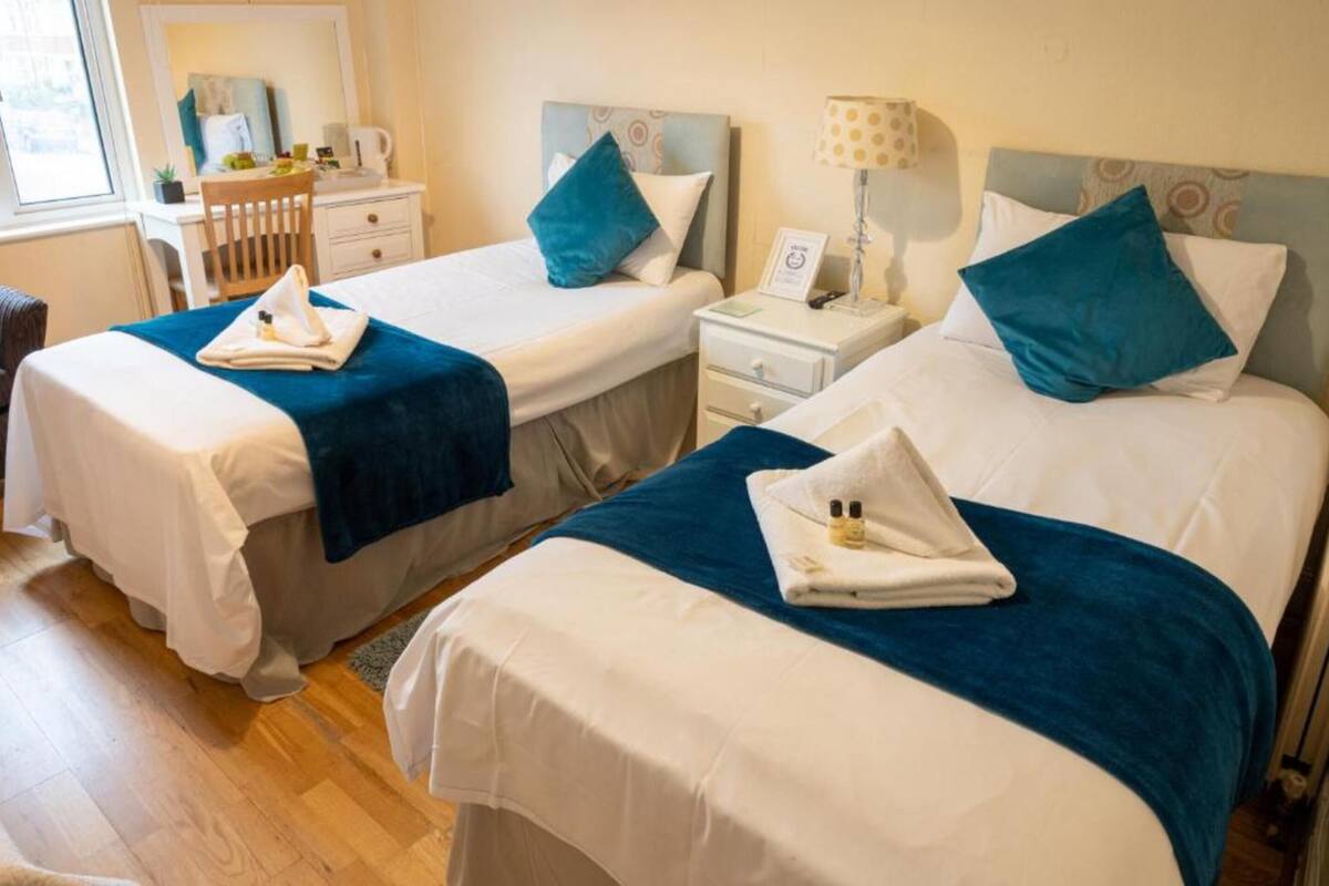 Camera d'albergo con due letti, ciascuno con coperta blu e cuscino bianco. È presente un comodino con lampada e telefono.