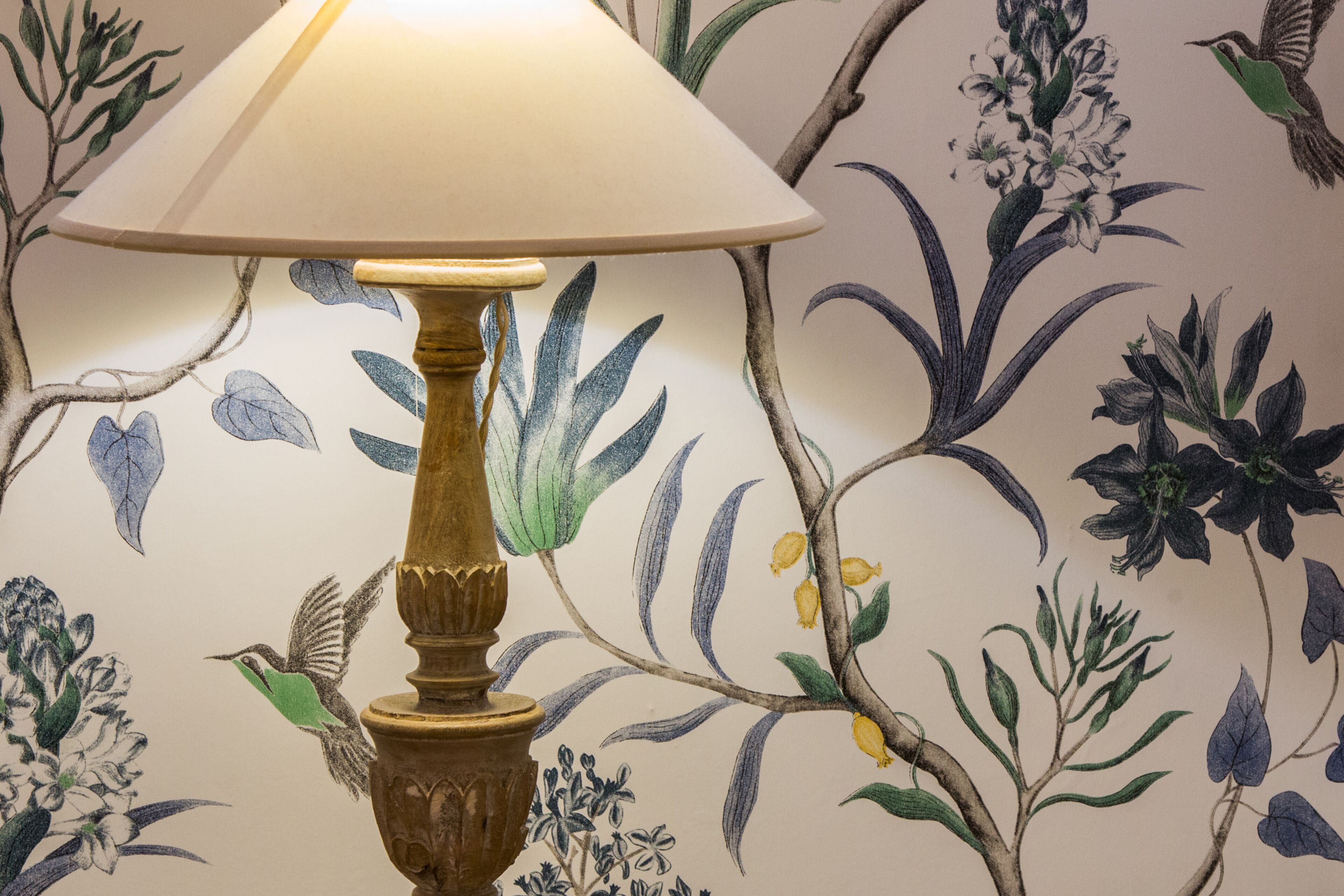 A lamp with a beige shade and a wooden base, placed on a wall with a floral wallpaper design.