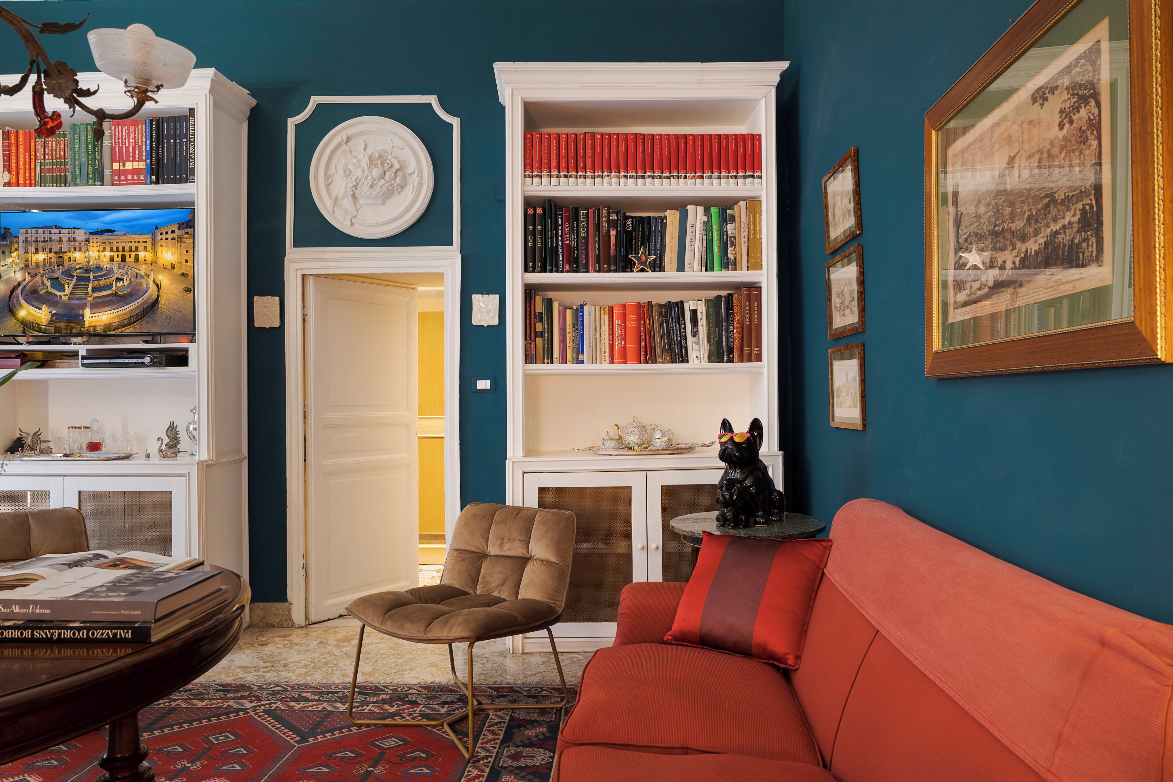 Een kamer met een rode bank, een boekenkast, een stoel en een ingelijst kunstwerk aan de muur.