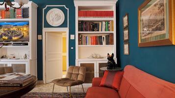 Een kamer met een rode bank, een boekenkast, een stoel en een ingelijst kunstwerk aan de muur.