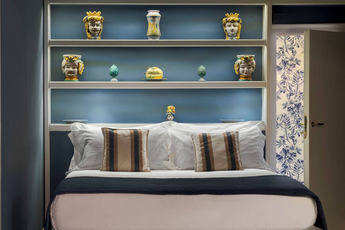 Uma cama com almofadas brancas e listradas, um cobreleite azul escuro e uma prateleira decorativa acima com diversos objetos de cerâmica.