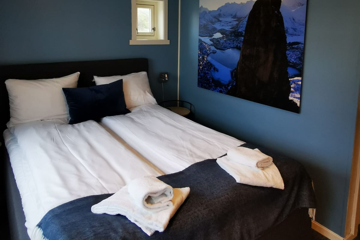 A bedroom with a bed, pillows, a nightstand, a wall-mounted window, and a painting of a mountain landscape.