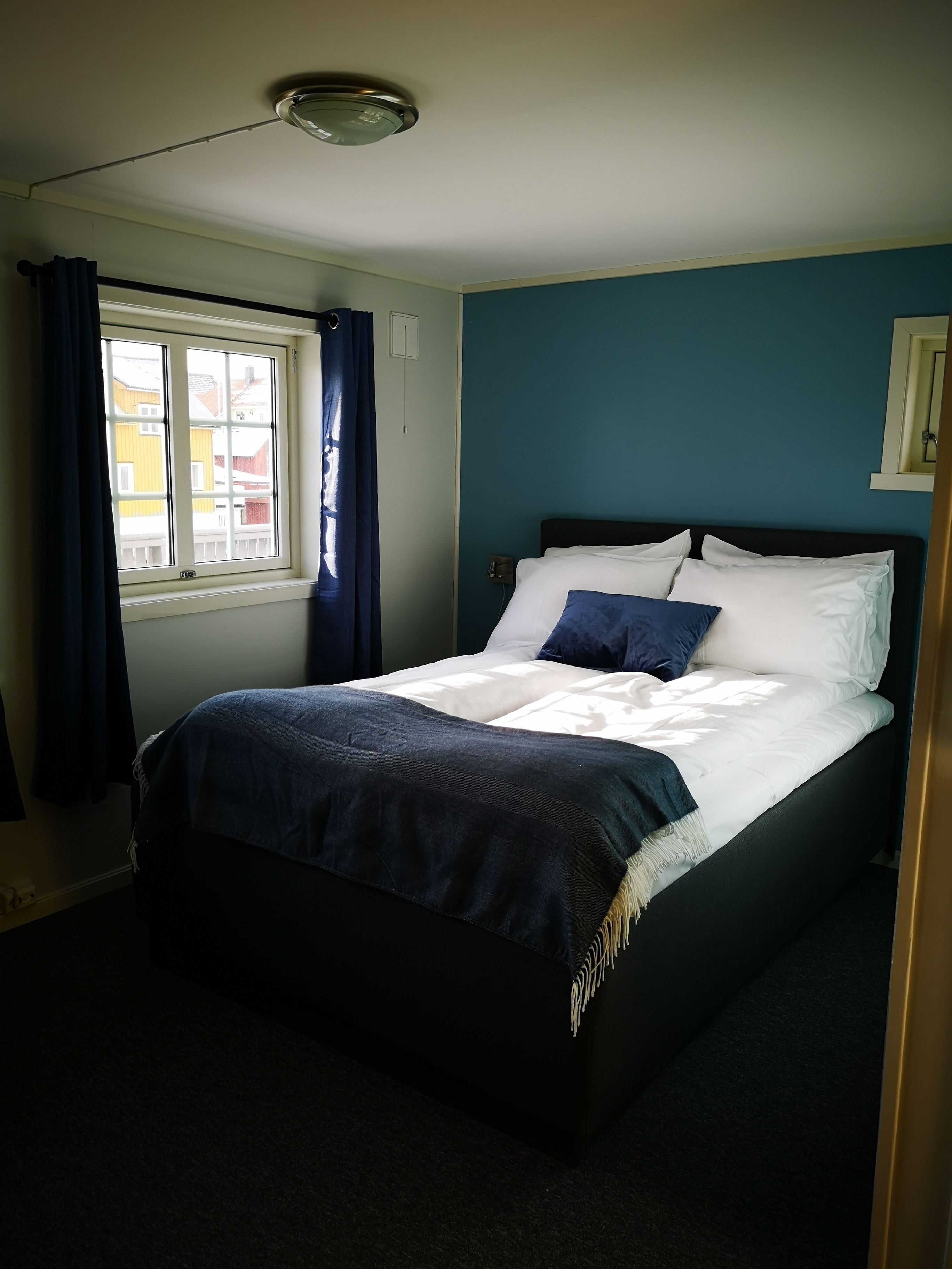 A bedroom with a bed, a window with curtains, a ceiling light, and a wall with a picture.